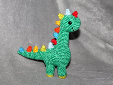 Gehäkelter grüner Amigurumi-Dino mit bunten Rückenzacken und gelben Fußenden auf grauem Hintergrund