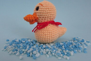 Gehäkelte Amigurumi-Ente in pfirsichfarbenem Garn mit orangefarbenem Schnabel und roter Schleife auf blau-weißen Kieselsteinen