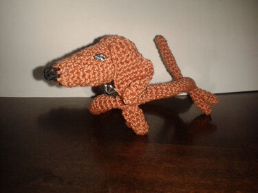 Braunes gehäkeltes Dackel-Amigurumi mit Nieten-Halsband auf dunkler Holzfläche.