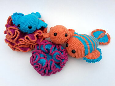 Gehäkelte Amigurumi-Fische und rüschenartige Korallen in Orange, Türkis und Magenta vor weißem Hintergrund