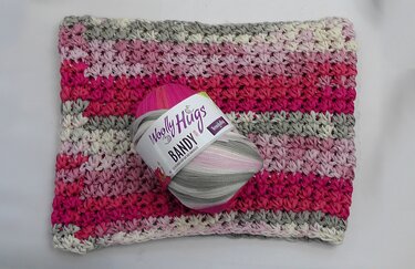 Gehäkelter Loopschal mit Streifen in Rosa, Pink, Grau und Creme, daneben ein Garnknäuel in passenden Farben.