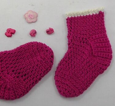 Zwei fuchsiafarbene gehäkelte Babysocken mit weißem Bündchen und rosa Dekorknöpfen auf hellem Hintergrund.