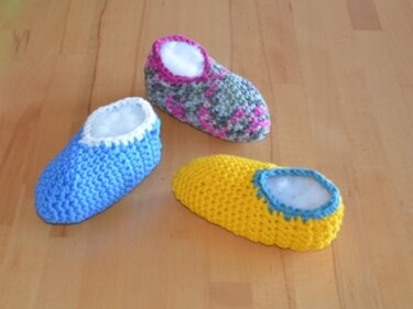 Drei gehäkelte Babyschuhe (blau, gelb, mehrfarbig) mit hellem Futter auf Holzuntergrund.