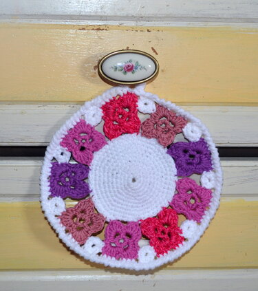 Runder handgehäkelter Topflappen mit quadratischen Blütenmotiven in Pink, Lila und Weiß, aufgehängt an einem Griff