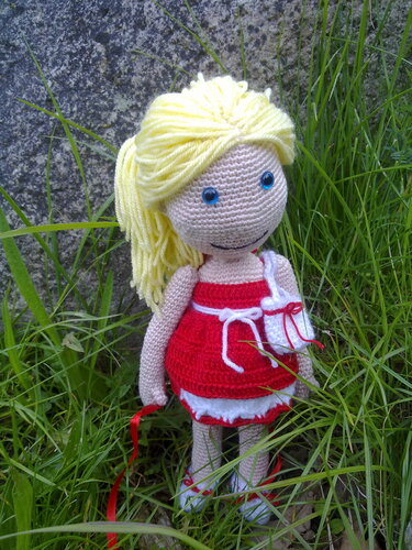 Gehäkelte Amigurumi-Puppe mit blondem Pferdeschwanz, rotem Kleid und kleiner weißer Tasche im Gras