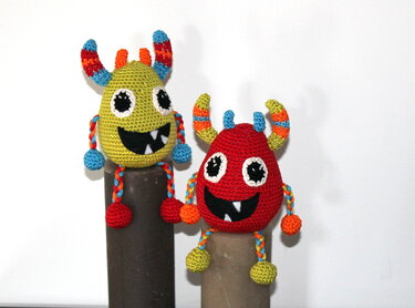 Zwei gehäkelte Amigurumi-Monster, links grün, rechts rot, mit gestreiften Hörnern und breitem Lächeln.