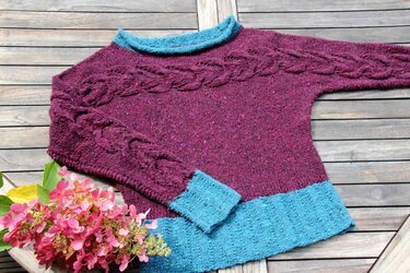 Damenpullover in Brombeerrot mit türkisfarbenem Kragen, Bündchen, Zopfmuster und rosa Blüten auf Holzuntergrund