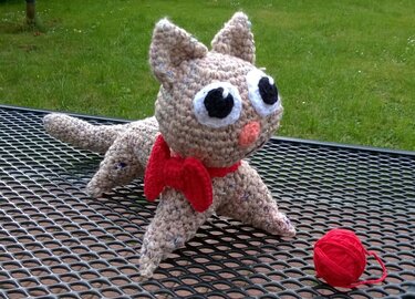 Gehäkeltes Amigurumi-Kätzchen mit großen Augen, roter Schleife und rotem Garnknäuel auf Metallgitter