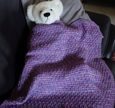 Weißer Teddybär unter lila gestrickter Babydecke mit Strukturmuster