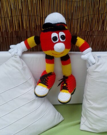 Gehäkelte Amigurumi-Figur in Schwarz-Rot-Gelb mit Mütze, Handschuhen und Turnschuhen auf weißem Sofa