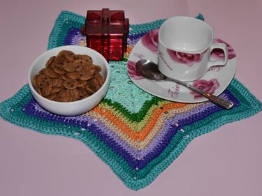 Mehrfarbiges gehäkeltes sternförmiges Deckchen mit Tasse, Untertasse, Löffel, Schale und kleinem Geschenk