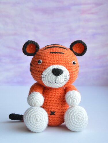 Front view of a crocheted orange amigurumi tiger showing stripes, white paws, and tail