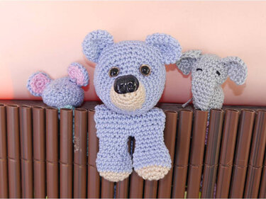 Kleines fliederfarbenes gehäkeltes Bären-Lesezeichen mit zwei Beinen und dunkler Nase auf braunem Lattenuntergrund, daneben zwei weitere gehäkelte Figuren