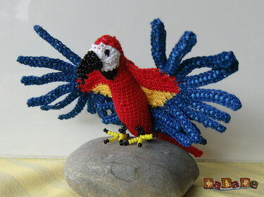 Gehäkelter Amigurumi-Papagei in Rot, Blau und Gelb mit perlenbesetzten Flügeln auf grauem Stein.
