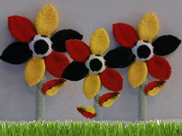 Drei gehäkelte Blumen in Schwarz, Rot und Gelb mit weiß-schwarzen Kernen und grünen Stielen vor hellem Hintergrund.