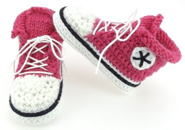 Nahaufnahme zweier gehäkelter Babyschuhe in Pink, Weiß und Schwarz mit Schnürsenkeln und rundem Seitenemblem