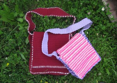 Zwei gehäkelte Umhängetaschen: burgunderrote Tasche mit weißer Kante und rosa-weiß gestreifte Tasche mit lila Träger auf Gras