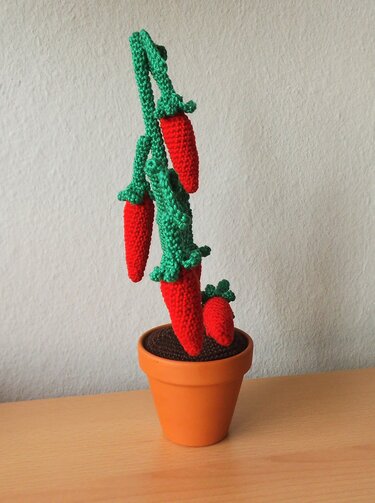 Gehäkelte rote Chilipflanze mit grünen Stielen in einem Terrakottatopf auf einer Holzoberfläche.