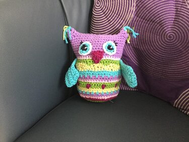 Farbig gehäkelte Eule mit violettem Kopf, gestreiftem Körper und türkisfarbenen Flügeln auf einem Sofa neben einem gemusterten Kissen