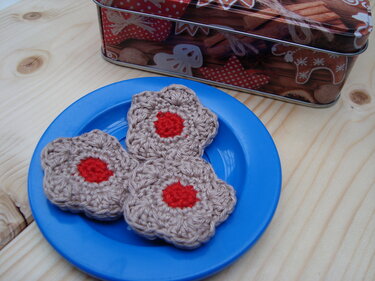 Drei gehäkelte Plätzchen in Beige mit roter Mitte auf einem blauen Teller neben einer weihnachtlichen Keksdose auf Holzuntergrund.