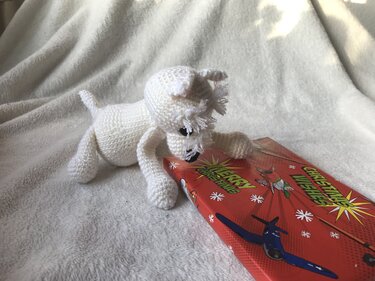Niedlicher gehäkelter weißer Amigurumi Hund mit Fransen am Kopf neben roter Geschenkbox