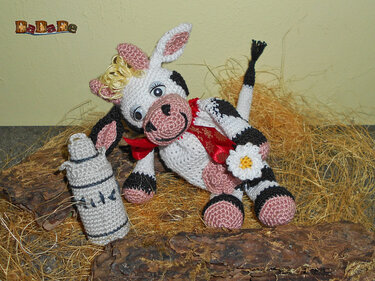 Gehäkelte schwarz-weiße Amigurumi-Kuh mit roter Schleife und Blume neben gehäkelter Flasche auf Stroh