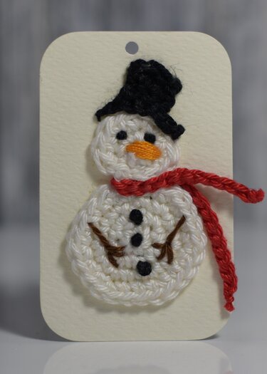 Gehäkelter Schneemann-Anhänger mit schwarzem Hut und rotem Schal auf beigem Anhänger