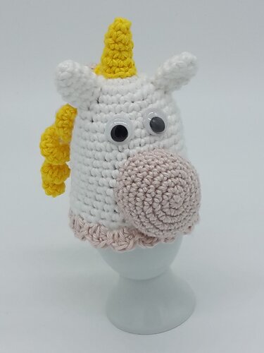 Gehäkelter Einhorn-Eierwärmer in Weiß mit gelber Mähne und Horn, rosa Schnauze und Wackelaugen auf weißem Eierbecher.