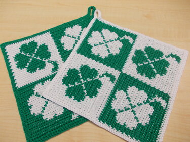 Zwei grün-weiße gehäkelte Topflappen mit Kleeblattmotiven auf Holzuntergrund.