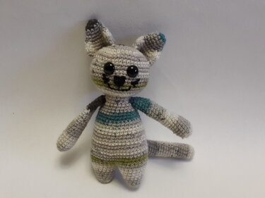 Gehäkelte Amigurumi-Katze in Grau mit türkisen und hellgrünen Streifen, großen schwarzen Augen und gesticktem Gesicht.