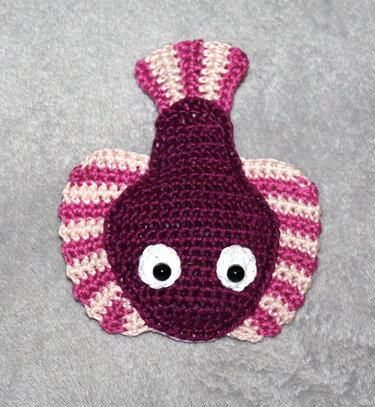 Flaches gehäkeltes Amigurumi-Fischmotiv in Dunkelviolett mit pink-rosa gestreiften Flossen und weißen Augen auf grauem Hintergrund