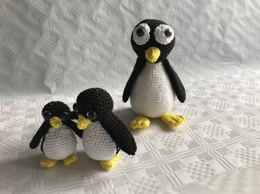 Drei gehäkelte Amigurumi‑Pinguine (zwei kleine, ein großer) auf weißem, gemustertem Stoff