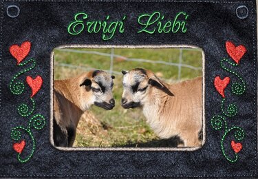Bestickter Rahmen mit zwei Lämmern, roten Herzen, grünen Ranken und dem Schriftzug Ewig' Liebi