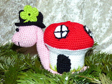 Rosa gehäkelte Schnecke mit schwarzem Zylinder, grünem Kleeblatt und rotem Pilzhaus-Schneckenhaus auf Tannennadeln.