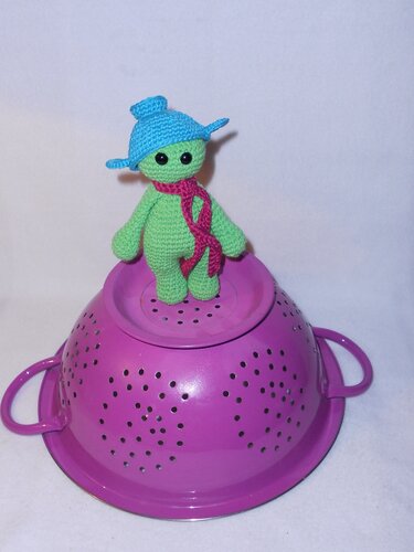 Gehäkeltes grünes Amigurumi mit blauem Hut und pinkem Schal auf lila Sieb