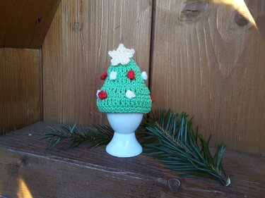 Gehäkelter grüner Eierwärmer in Form eines Weihnachtsbaums mit Stern und roten sowie weißen Verzierungen auf einem weißen Eierbecher