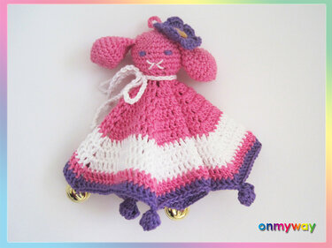 Gehäkeltes Häschen‑Schmusetuch in Rosa, Weiß und Lila mit kleinen Glöckchen am Saum auf hellem Hintergrund.