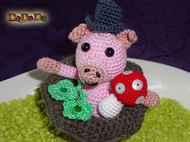 Gehäkeltes rosa Amigurumi-Glücksschwein mit kleinem Hut, rotem Pilz und grünen Kleeblättern in braunem Nest