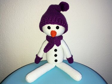 Gehäkelter weißer Schneemann mit lila Mütze und Schal auf blauem Untergrund
