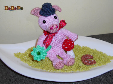 Rosa gehäkeltes Amigurumi-Glücksschwein mit blauem Hut, roter Schleife und gehäkelten Glückssymbolen in weißer Schale