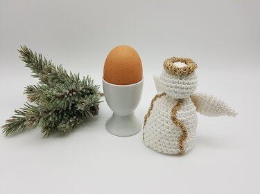 Weißer gehäkelter Engel-Eierwärmer mit goldfarbenem Heiligenschein neben Ei im weißen Eierbecher und Tannenzweig.