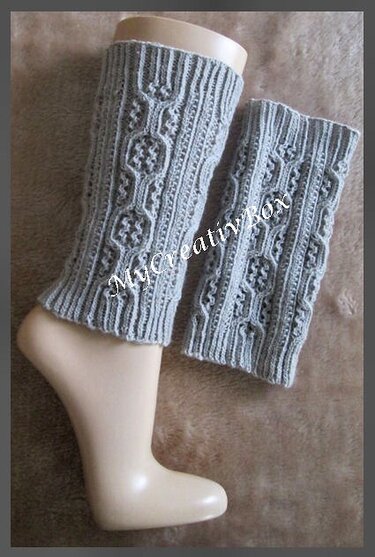 Zwei hellgraue gestrickte Kinder-Beinstulpen mit Zopfmuster, eine auf einem Beinmodell, die andere daneben liegend.
