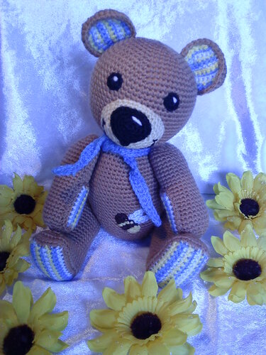 Gehäkelter brauner Teddybär mit blau-gelb gestreiften Ohren und Pfoten, blauem Schal und Bienen-Applikation vor gelben Sonnenblumen