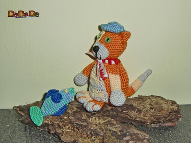 Gehäkelte orange-graue Amigurumi-Katze mit blauem Käppi und rot-weißem Schal neben gehäkeltem blau-grünem Fisch auf Rindenstück