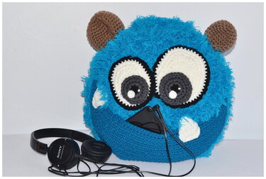Blaues gehäkeltes Monsterkissen mit großen Augen, Ohren und Vordertasche, darin ein Smartphone; Kopfhörer liegen daneben