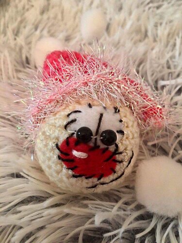 Runder gehäkelter Amigurumi-Schlüsselanhänger in Beige mit schwarzen Knopfaugen, rotem Mund und pinkem Glitzergarn auf weißem, flauschigem Stoff
