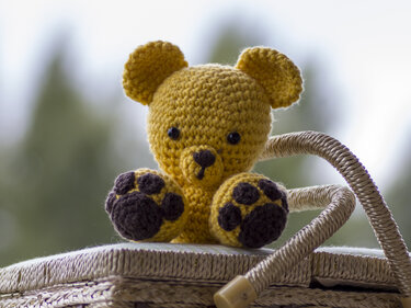 Kleiner gelber gehäkelter Amigurumi-Teddybär mit dunkelbraunen Tatzen auf einem geflochtenen Korb