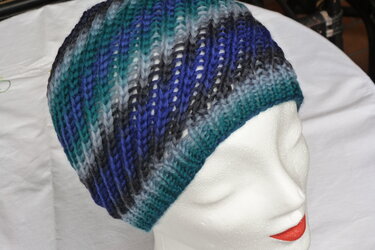 Gestrickte Beanie-Mütze in Blau-, Türkis- und Grautönen auf weißem Schaufensterkopf. Vertikales Rippenmuster erkennbar.
