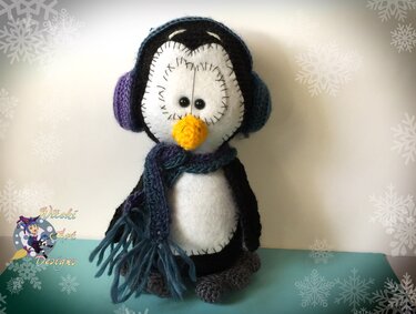 Gehäkelter Amigurumi-Pinguin mit lila-blauen Ohrenschützern und Schal vor hellem Hintergrund mit Schneeflocken.