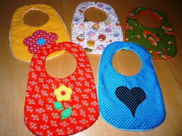 Fünf bunte Baby-Lätzchen mit unterschiedlichen Stoffmustern und applizierten Motiven auf einem Holztisch.